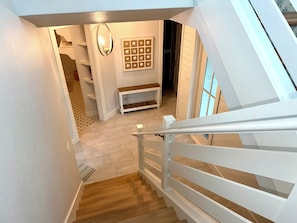 Una escalera con barandillas blancas que conduce a una habitación con piso de madera, un aplique en la pared y un cuadro enmarcado.