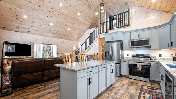 A spacious living area with a large island, a sofa, and a staircase leading to an upper level. - Quimby Cabin | New Build | Snowmobile Access (Rangeley)