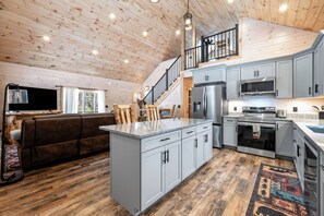 A spacious living area with a large island, a sofa, and a staircase leading to an upper level. - Quimby Cabin | New Build | Snowmobile Access (Rangeley)