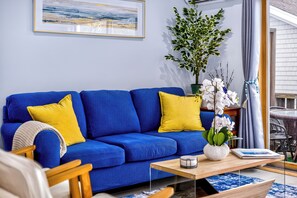 Ein blaues Sofa mit gelben Kissen, ein Holztisch, eine Topfpflanze und ein gerahmtes Bild an der Wand.