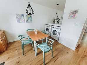 Una cocina con una mesa de madera y sillas color turquesa, una lavadora y dos cuadros abstractos en la pared.