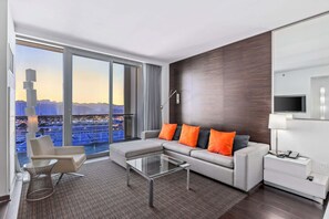 A modern hotel room with a large window offering a city view, a sofa with orange cushions, a glass coffee table, and a bedside table with a lamp. - Incredible Views 51st Floor High-Rise Studio -Balcony (Las Vegas)