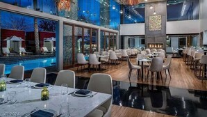 A modern dining area with a pool view, large windows, and a central fireplace. - Incredible Views 51st Floor High-Rise Studio -Balcony (Las Vegas)