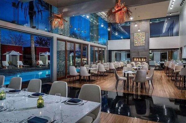 A modern dining area with a pool view, large windows, and a central fireplace. - Incredible Views 51st Floor High-Rise Studio -Balcony (Las Vegas)