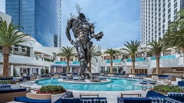 Zona de piscina con una estatua, palmeras y edificios modernos al fondo.