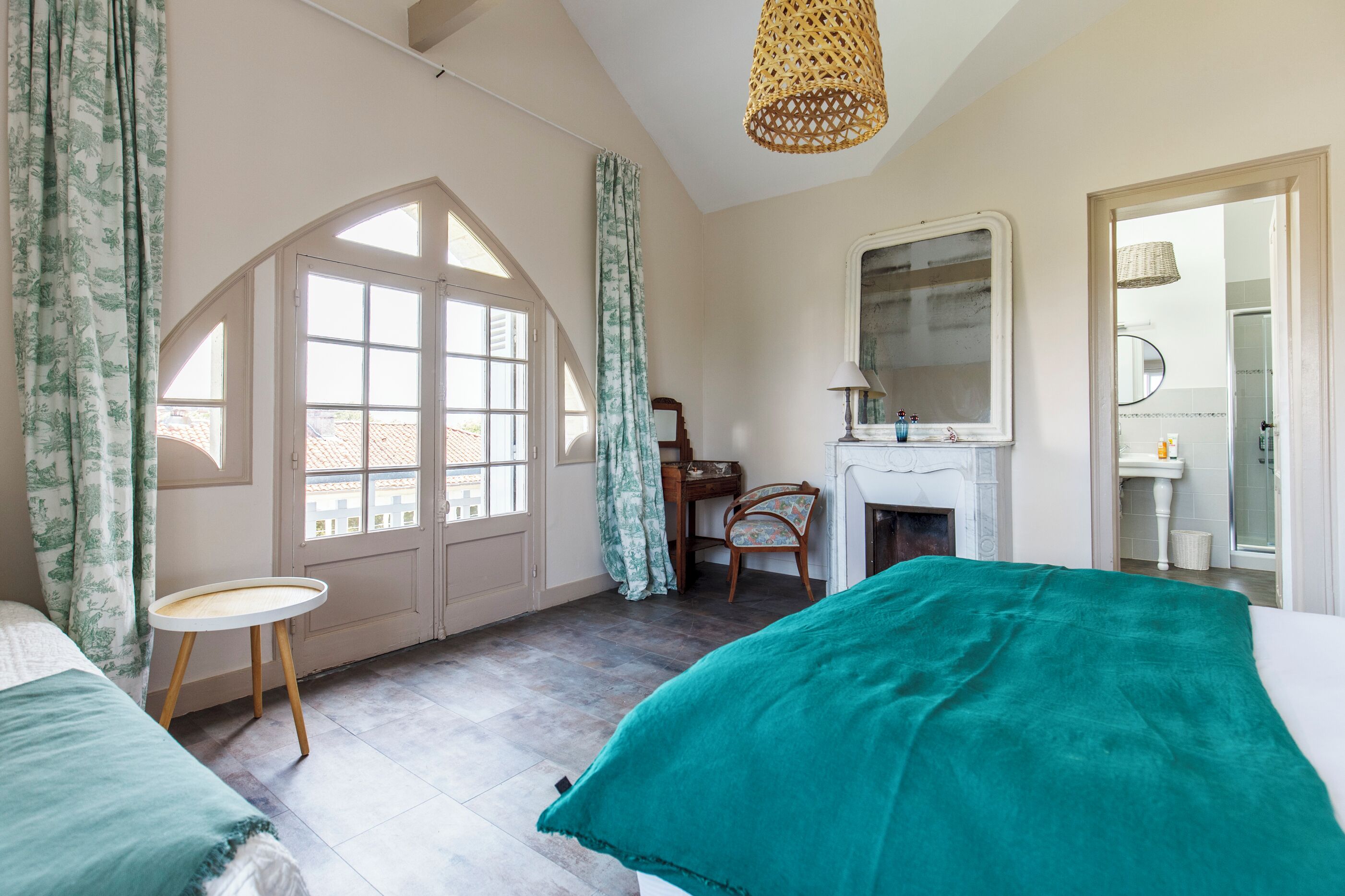 Un dormitorio con cama, una mesita, una silla, chimenea y ventana con cortinas.