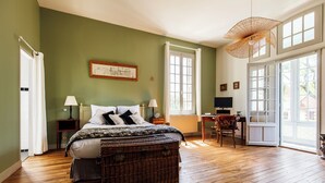 A bedroom with a large bed, a desk, a chair, a wicker chest, and a large window.
