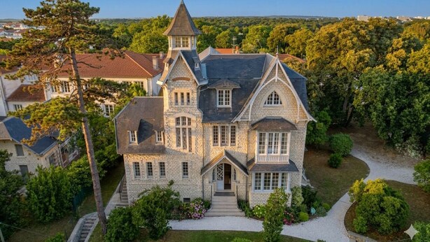 A large, historic stone house with a prominent tower, surrounded by lush greenery and a well-maintained lawn.