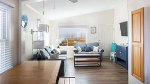 Ein Wohnzimmer mit Sofa, Couchtisch und Lampe; ein Essbereich mit Tisch; ein Deckenventilator; ein Fenster mit Jalousien; eine Tür und ein Blick auf ein Gebäude.