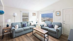 Ein Wohnzimmer mit zwei grauen Sofas, einem Holztisch und Blick auf das Meer durch die Fenster.