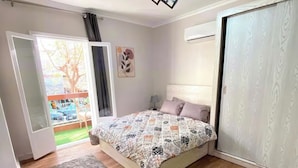 Ein Schlafzimmer mit Bett, zwei Teppichen, einer Deckenleuchte, einer Tür und einem Fenster mit Blick auf einen Baum und ein Gebäude.