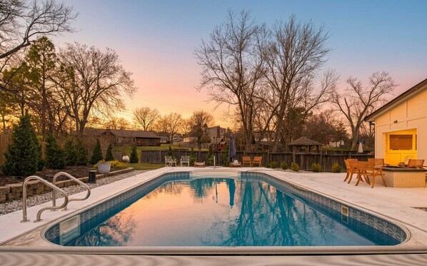 A rectangular swimming pool with a tiled edge, surrounded by a deck with chairs and a gazebo.