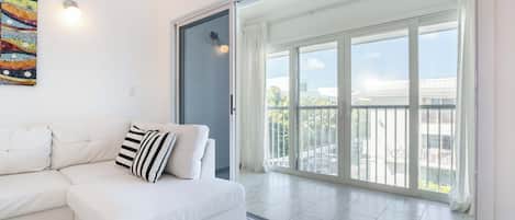 Sofá branco com almofadas listradas, pintura abstrata colorida e uma grande porta de vidro deslizante que dá para uma varanda com vista para edifícios e áreas verdes.