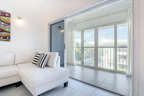 En hvid sofa med stribede puder, et farverigt abstrakt billede og en stor skydedør, der fører ud til en balkon med udsigt over bygninger og grønt.