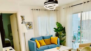Een moderne woonkamer met een blauwe bank, gele kussens, een beige fauteuil, een witte salontafel en een plafondventilator.