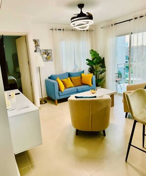 A modern living room with a blue sofa, yellow cushions, a beige armchair, a white coffee table, and a ceiling fan. - Cozy 2BR Retreat w/ Pool, WiFi, Parking – Minutes to Beach (Punta Cana)