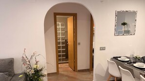 Ein modernes Wohnzimmer mit grauem Sofa, einem Essbereich mit Tisch für zwei Personen und einem Flur, der zu einem Badezimmer mit Glasdusche führt.