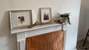 Une cheminée surmontée d’une étagère décorative, avec deux tableaux encadrés, une branche décorative et une plante en pot.