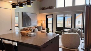 A modern kitchen with a central island, a dining area with chairs, a sofa, and a large window with a view. - Timberline Hydeaway at Ascent (Canmore)
