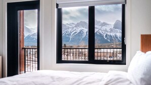 A bedroom with a bed, a window with a view of snow-capped mountains, and a balcony with a railing. - Timberline Hydeaway at Ascent (Canmore)