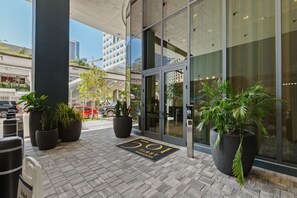 Entrada de hotel con puerta de vidrio, plantas en macetas, felpudo y aparcamiento al fondo.