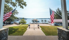 A view from a porch with two American flags, a pathway leading to a dock, and a body of water in the background.