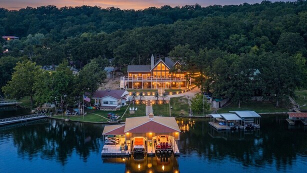 A large, multi-story house with a dock extending into a lake, surrounded by dense forest. - Honeysuckle House (Gravois Mills)