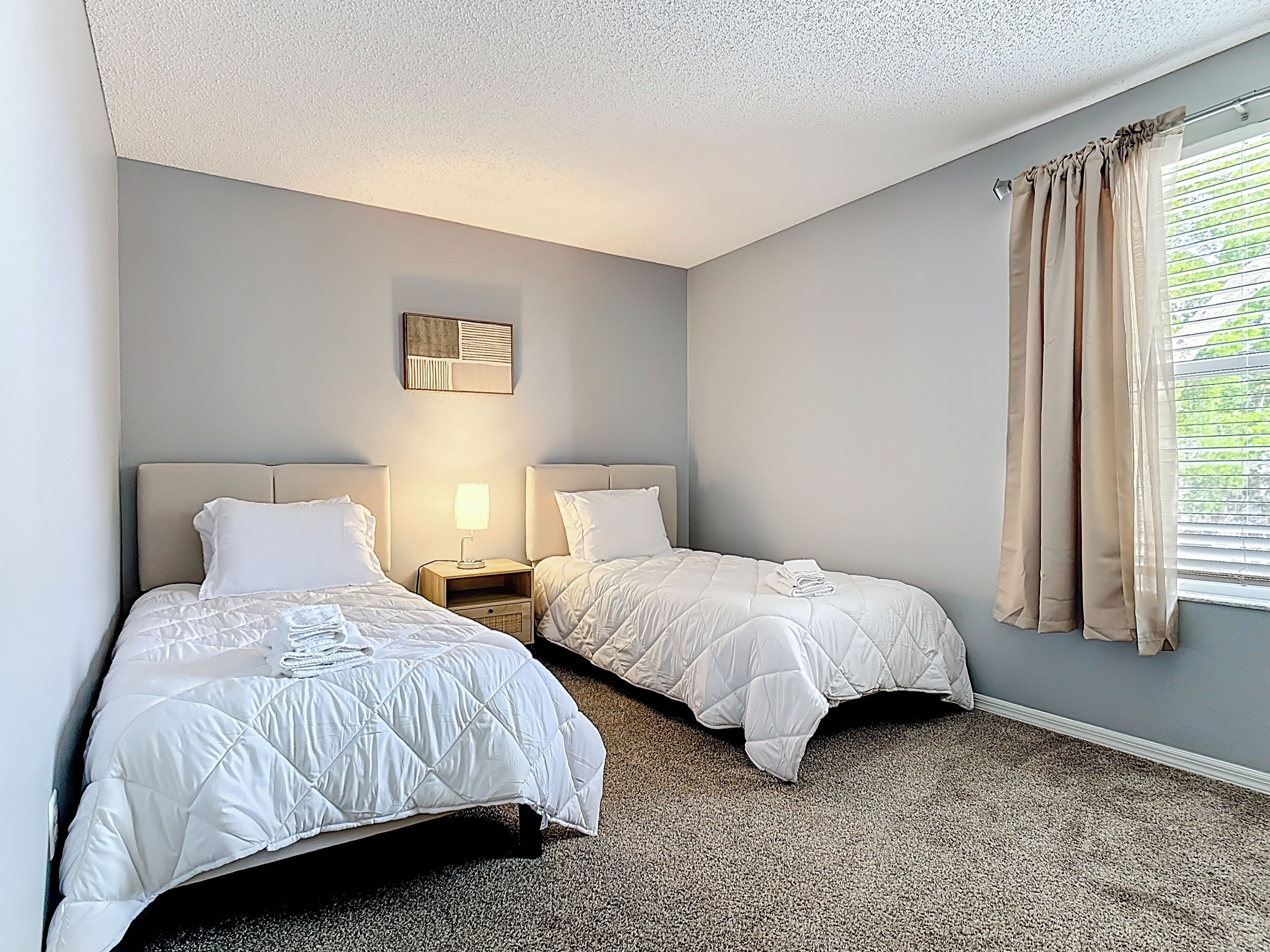 A hotel room with two beds, a nightstand with a lamp, a wall clock, and a window with curtains.