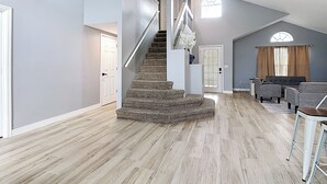 Amplia sala de estar con piso de madera, una escalera, una zona de estar con sofá y sillones, y un ventanal con cortinas.