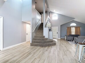 Ein geräumiger Wohnbereich mit Holzboden, einer Treppe, einer Sitzecke mit Sofa und Sesseln und einem fenster mit Vorhängen.