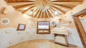 A room with a wooden ceiling, a bed with pillows, a window with a view, and a wall with circular cutouts.