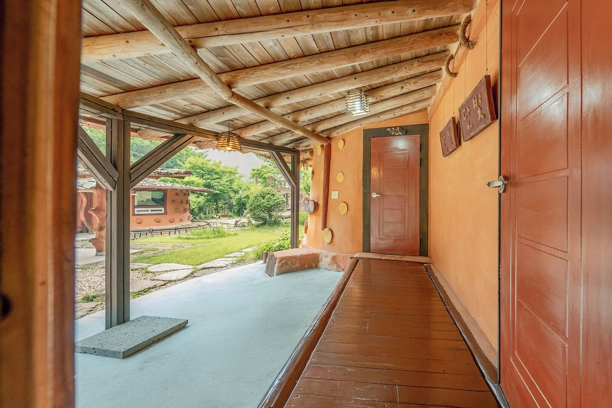 A wooden hallway with a concrete floor, a red door, and a view of a garden with a wooden structure.
