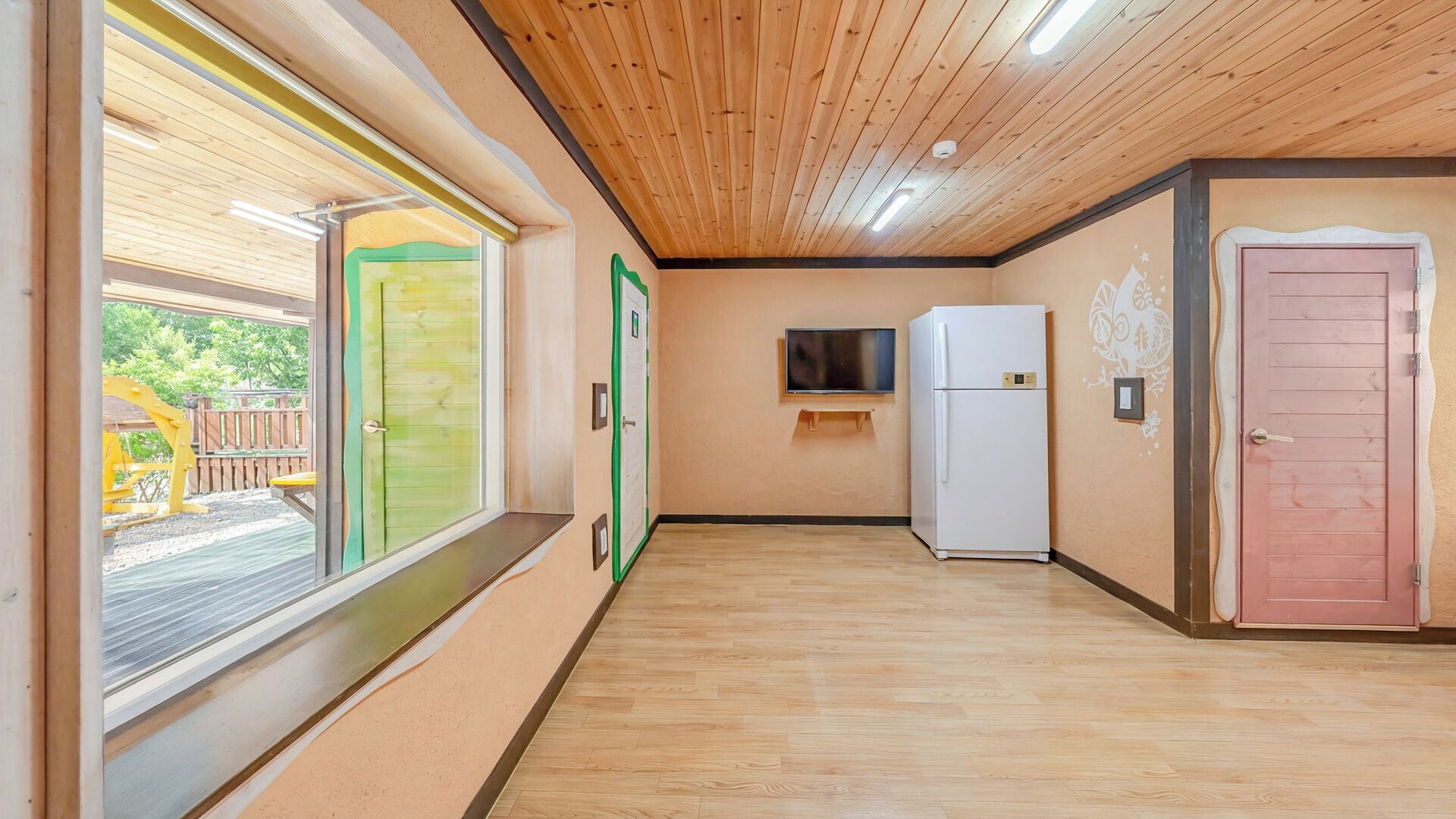 Ein Zimmer mit Holzboden, einem an der Wand montierten Fernseher, einem Kühlschrank und einer Tür mit einem besonderen Design.