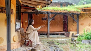 A person sitting on a bench under a wooden structure with a thatched roof, surrounded by a rustic outdoor setting. - Mungyeong Between Mountains Pet Pension (Mungyeong)