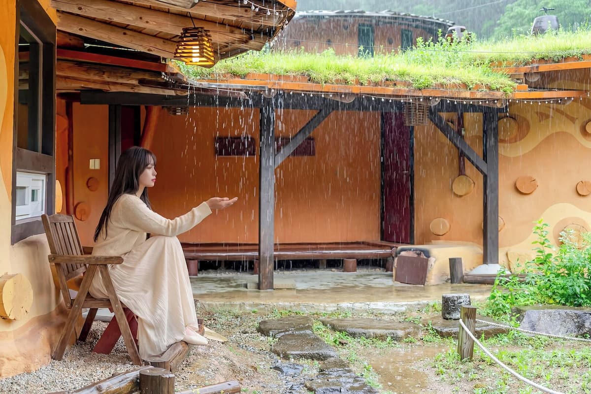 A person sitting on a bench under a wooden structure with a thatched roof, surrounded by a rustic outdoor setting.