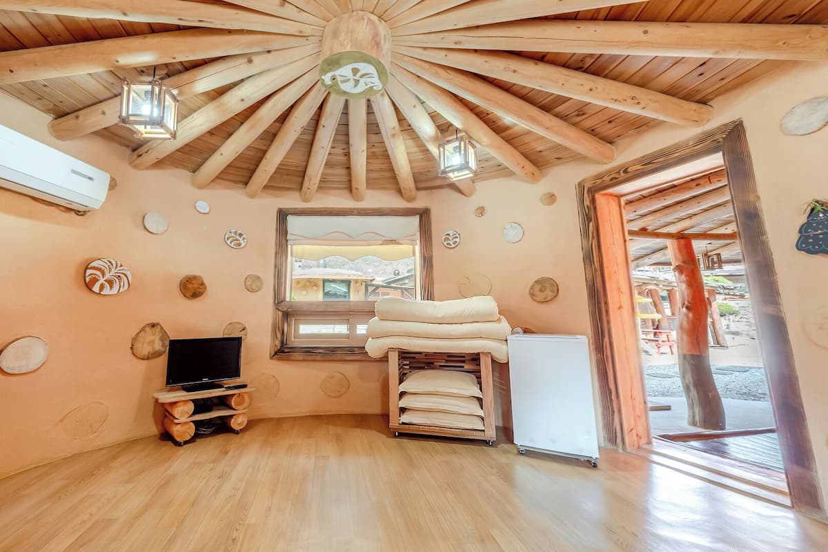 A room with a wooden ceiling, a television, a bed with pillows, and a window with a view.
