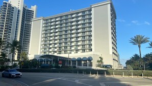 A multi-story hotel with a modern design, featuring a central entrance with a covered area, surrounded by palm trees and a clear sky. - Hotel Sunny Isles (Sunny Isles Beach)