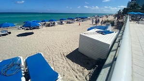 A beach with blue umbrellas, lounge chairs, and a building in the background. - Hotel Sunny Isles (Sunny Isles Beach)