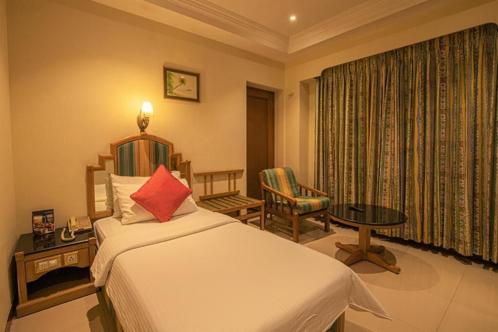 A hotel room with a bed, a chair, a small table, a nightstand, a lamp, a framed picture, and a curtain.