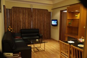 Una sala de estar con un sofá negro, una mesa de centro de cristal, una mesa de comedor de madera con sillas, un televisor y un mueble de madera con una planta.