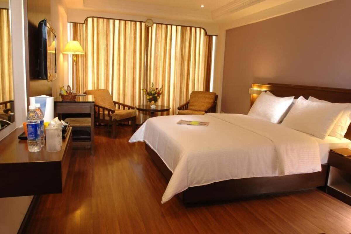 A hotel room with a large bed, a desk with a lamp, a chair, a small table with a vase, and a TV mounted on the wall.