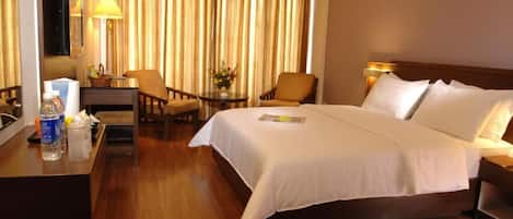 Habitación de hotel con una cama grande, un escritorio con lámpara, una silla, una mesita con un florero y un televisor montado en la pared.