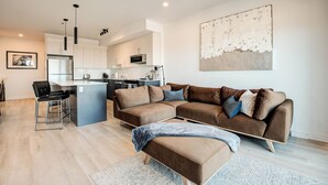 Een moderne woonkamer met een bruine modulaire bank, een keuken op de achtergrond en een groot abstract schilderij aan de muur.