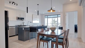 Een moderne keuken met een centraal eiland, een eettafel met stoelen, hanglampen en een groot raam dat voor daglicht zorgt.