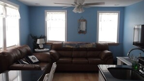 A living room with a brown leather sofa, a glass coffee table, a ceiling fan, and a kitchen area with a sink and cabinets. - Beautiful 1-bedroom condo across the street from the beach w/ AC (Brigantine)
