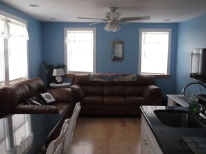 A living room with a brown leather sofa, a glass coffee table, a ceiling fan, and a kitchen area with a sink and cabinets. - Beautiful 1-bedroom condo across the street from the beach w/ AC (Brigantine)