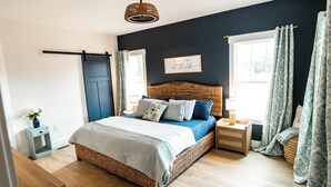 Quarto com cama, mesas de cabeceira, prateleira na parede, porta e janela com cortinas.