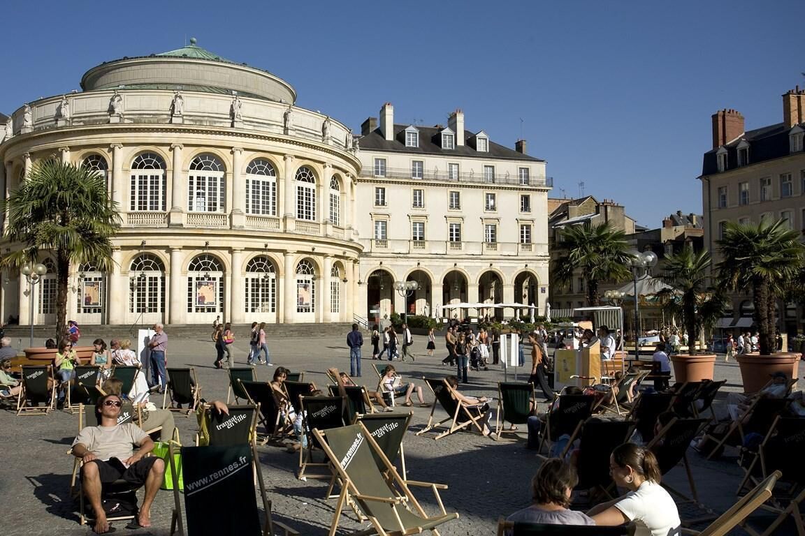 A public square with a historic building featuring arched windows and a dome. People are sitting on chairs, and there are palm trees around.