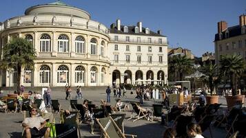 A public square with a historic building featuring arched windows and a dome. People are sitting on chairs, and there are palm trees around.