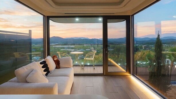 A modern living room with a sofa, two chairs, and a small table. A large sliding glass door leads to a balcony with a view of mountains and a cityscape. - Gyeongju Holiday Space Pension (Gyeongju)